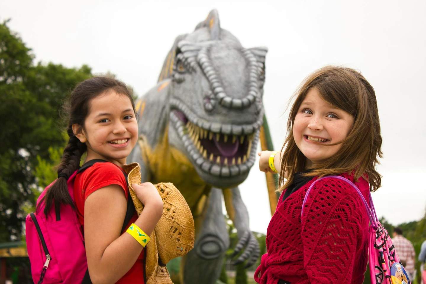 Two smiling girls with backpacks pose in front of a large dinosaur statue outdoors. One girl points at the dinosaur.
