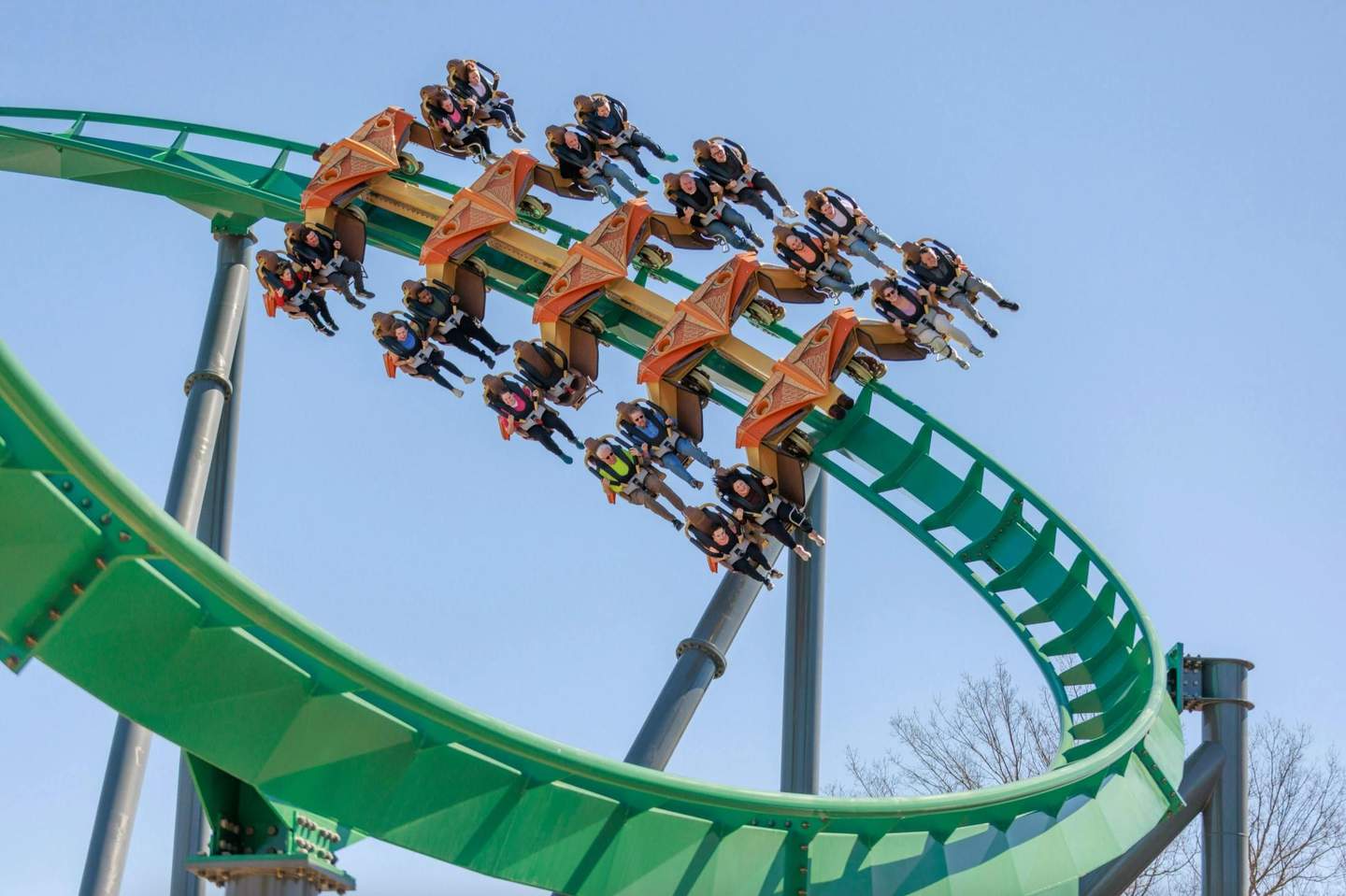 A roller coaster with green tracks and orange cars, carrying riders upside down on a loop under a clear blue sky.