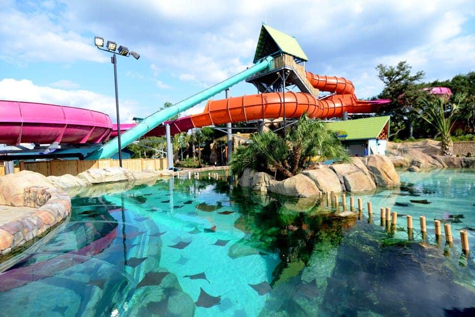 Water park with colorful slides, clear pools with stingrays, rocky landscaping, and palm trees under a partly cloudy sky.