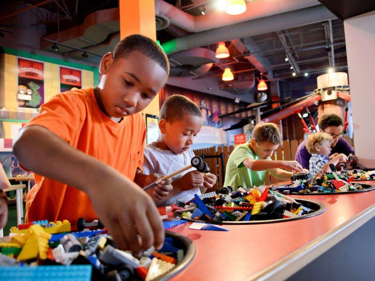 Children building with LEGO bricks at a play area, focused on their creations amid colorful scattered pieces on round tables.