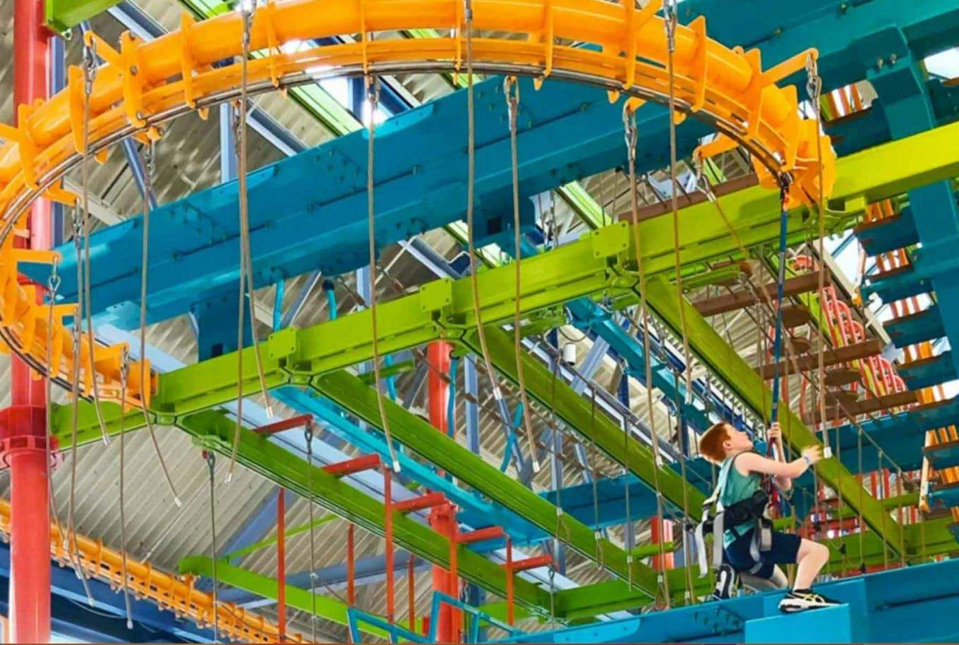 A young boy climbs a colorful indoor ropes course with blue, green, orange, and red structures and safety harnesses.