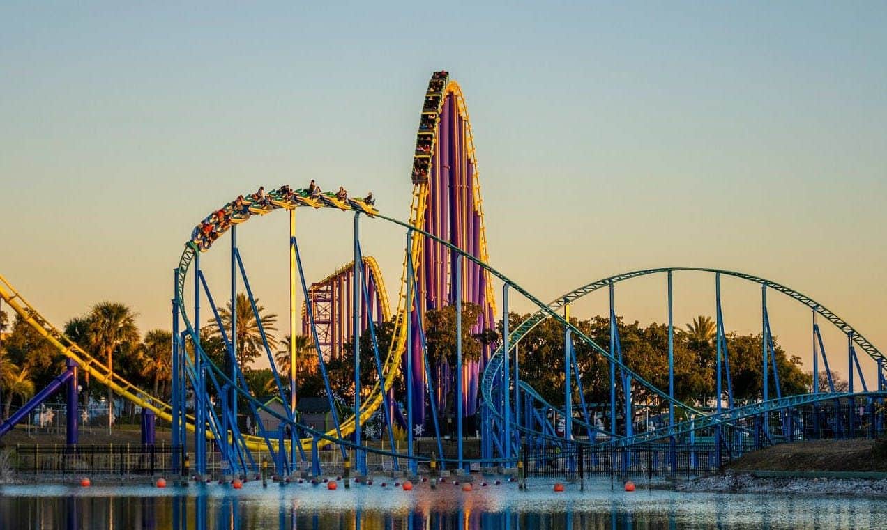 People riding a rollercoaster with blue and yellow tracks, looping over a body of water at sunset with trees in the background.