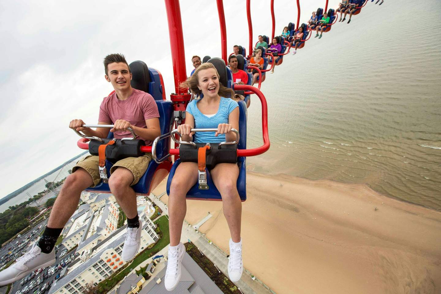 People enjoying a high swing ride overlooking a beach and buildings, smiling and holding onto the safety bars.