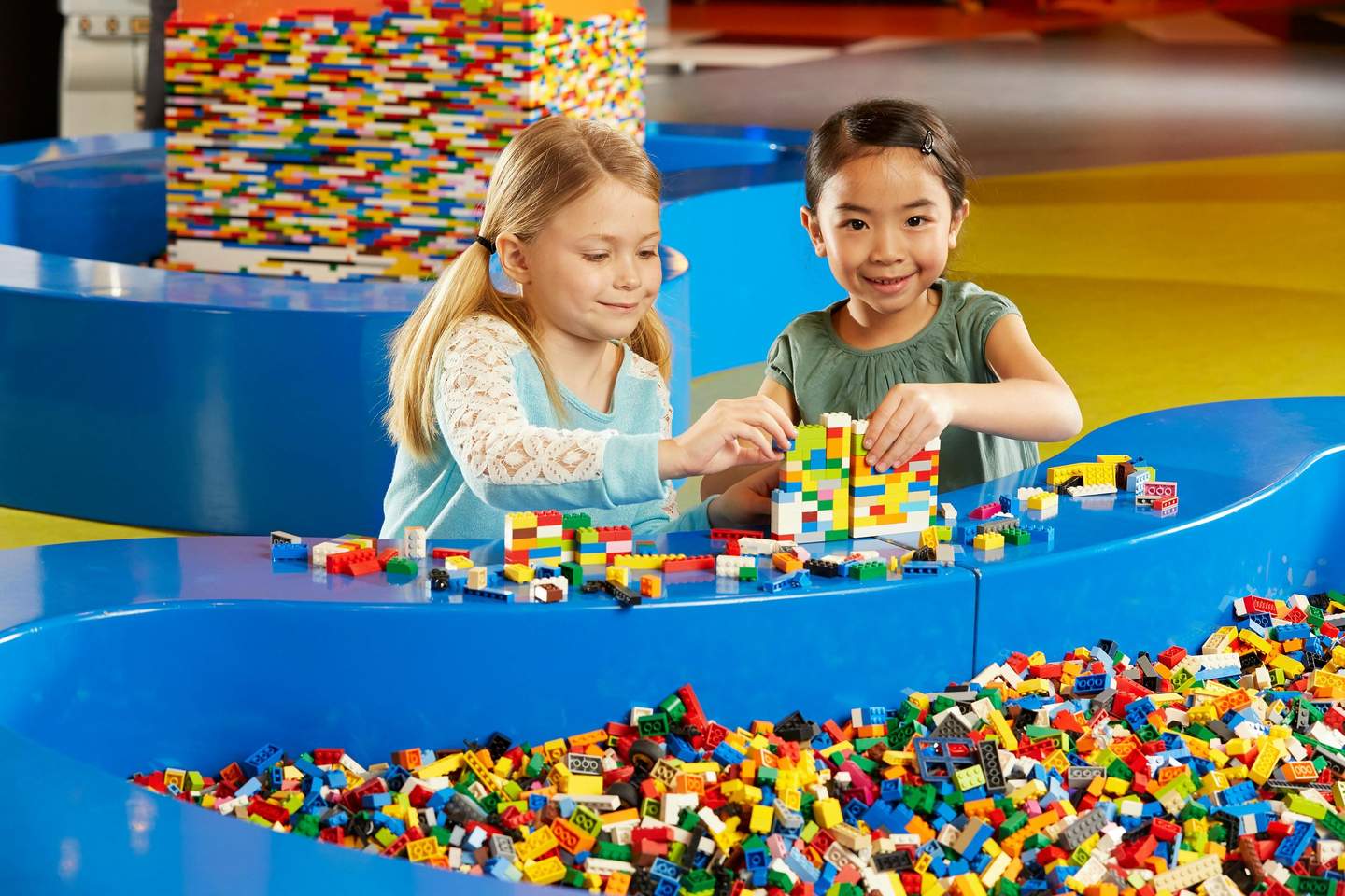 Two girls building structures with colorful LEGO bricks at a table filled with more LEGO pieces.