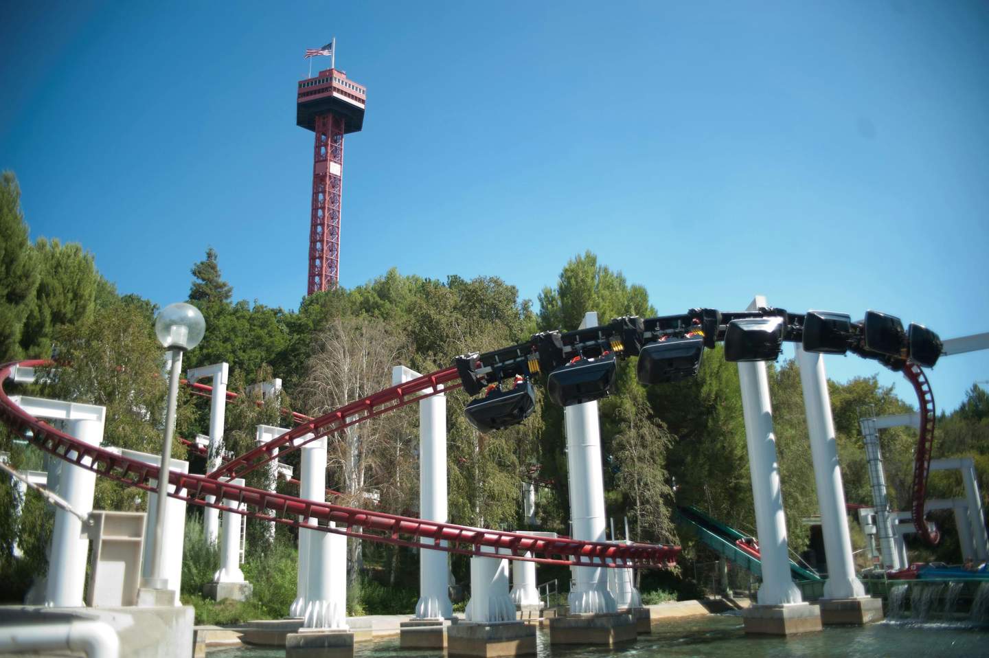 Six Flags Magic Mountain Park Tickets (València) A red rollercoaster track with suspended cars next to a tall red observation tower against a blue sky and green trees.