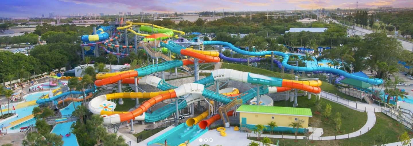 Large water park with numerous colorful water slides, pools, and a few people enjoying the attractions among trees and buildings.