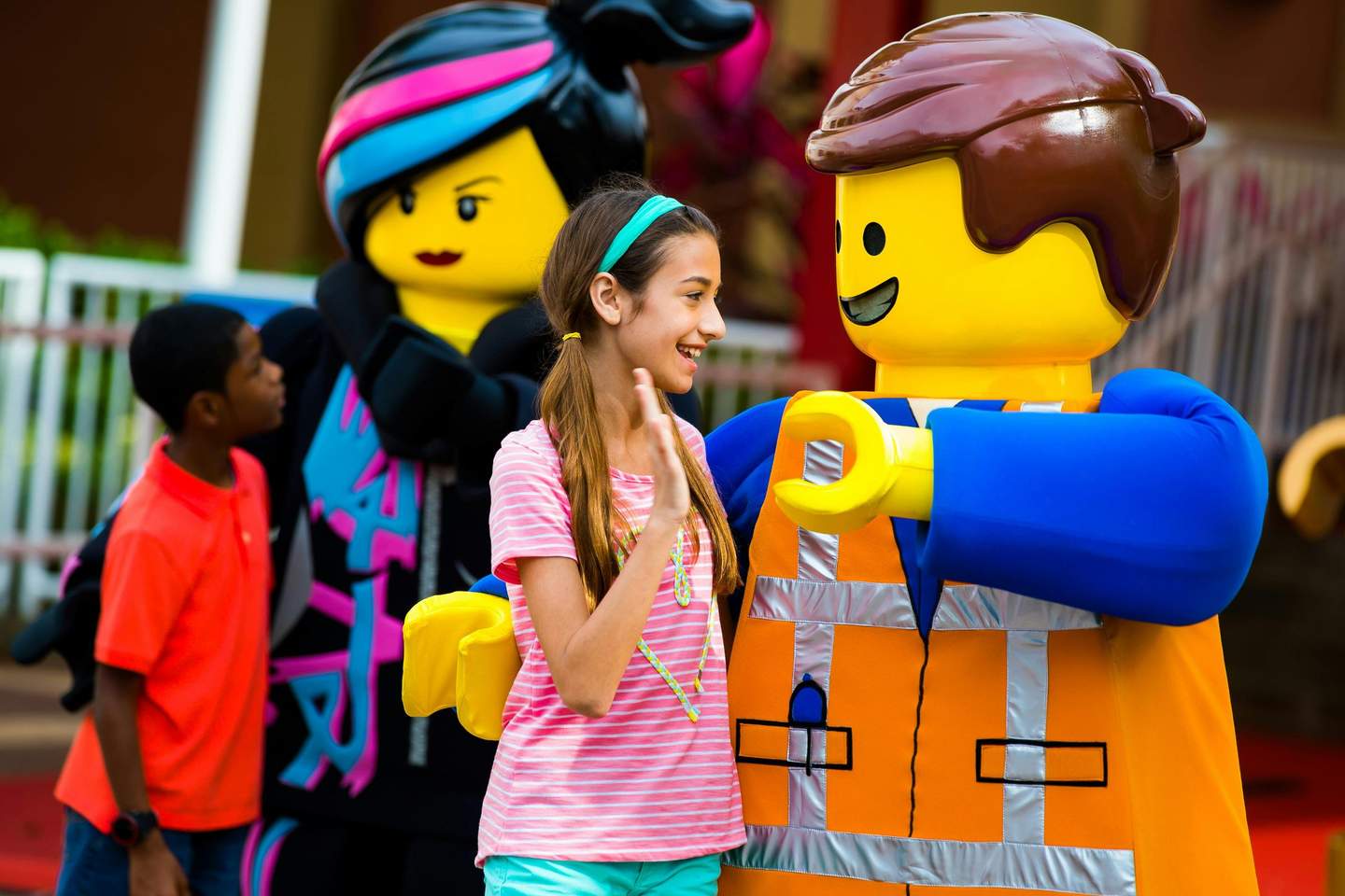 Two children interact with large costumed LEGO characters, one in an orange vest and the other with colorful hair, outdoors.