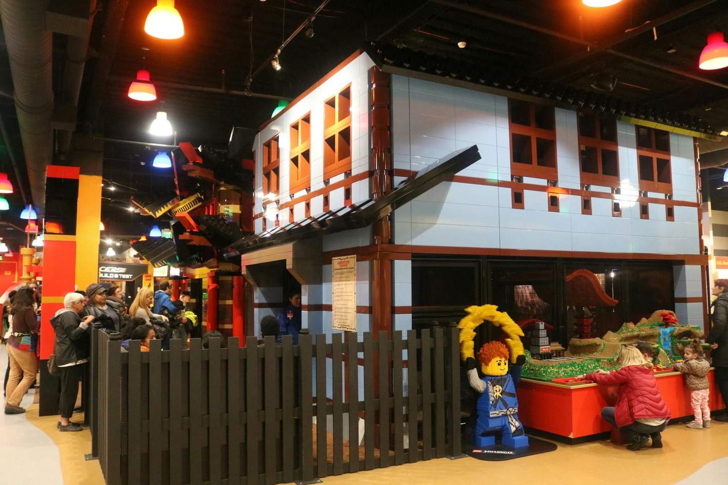 People exploring a brightly lit indoor Lego exhibition, featuring a large blue and brown Lego building and various Lego structures.