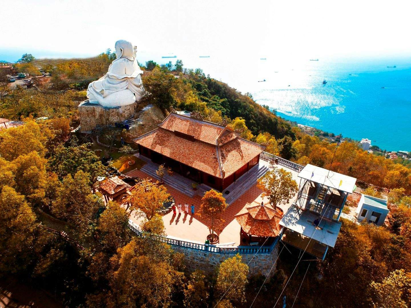 Aerial view of a Buddhist temple complex with a large white statue on a hill overlooking the sea, surrounded by trees.