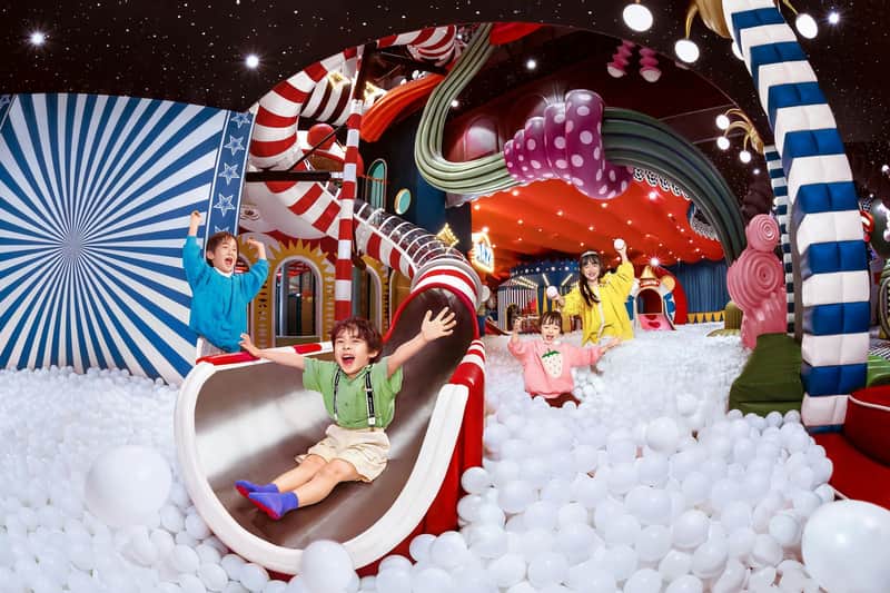 Children joyfully sliding down a slide into a ball pit in a colorful indoor playground with a whimsical design.
