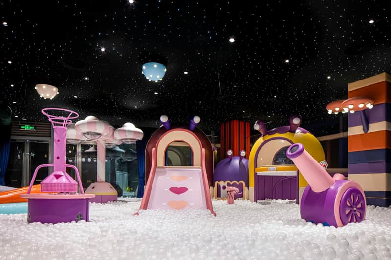 A colorful indoor playground with slides, structures shaped like snails, and a ball pit under a starry ceiling.