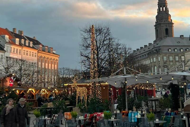 Copenhagen: Guided Christmas Tour + Tivoli Gardens Entry + Food & Drinks (Copenhagen) Outdoor seating area with decorated trees, string lights, and umbrellas. Buildings and a cloudy sky are visible in the background.