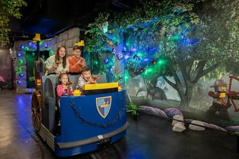 A family of four enjoys a ride in a themed cart at an indoor attraction with colorful decorations and lighting.