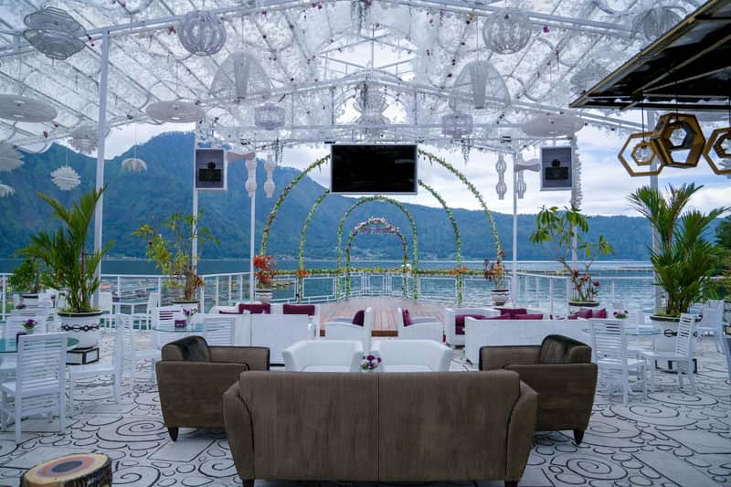 An outdoor seating area with sofas under a decorated canopy, overlooking a lake with mountain scenery in the background.