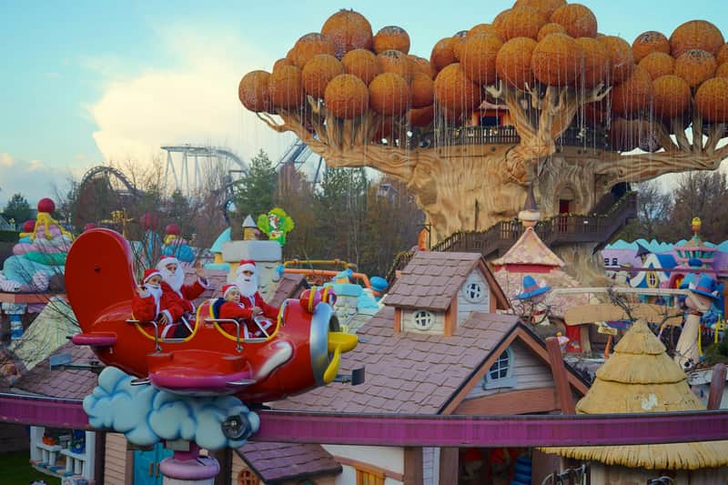 Santa Claus and helpers wave from a plane ride in an amusement park, featuring a large treehouse and whimsical buildings.