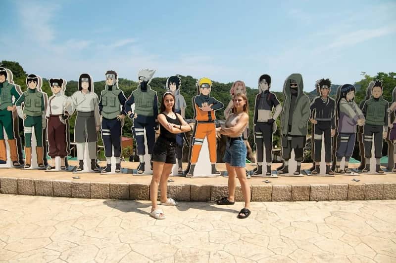 Nijigen No Mori Theme Park: NARUTO & BORUTO SHINOBI-ZATO VIP Journey Pass – Nijigen no Mori (Awaji) Two people pose in front of life-sized cardboard cutouts of anime characters outdoors on a sunny day.