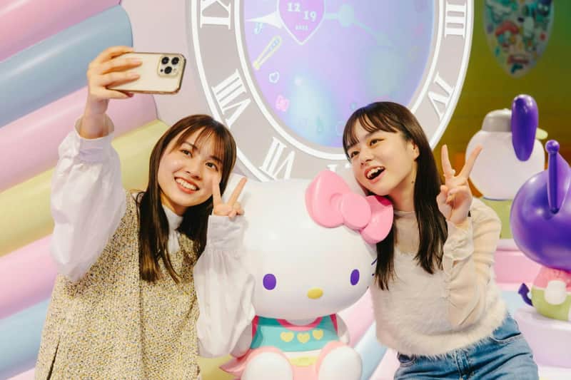 Two people posing with a Hello Kitty statue in a colorful setting with a clock-themed backdrop.