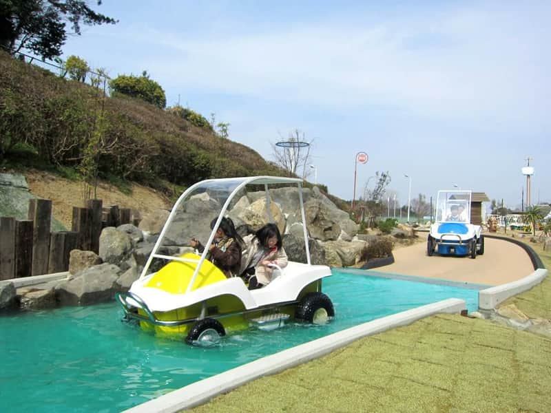 Two people riding a small amphibious vehicle in a shallow water channel, with another similar vehicle in the background.
