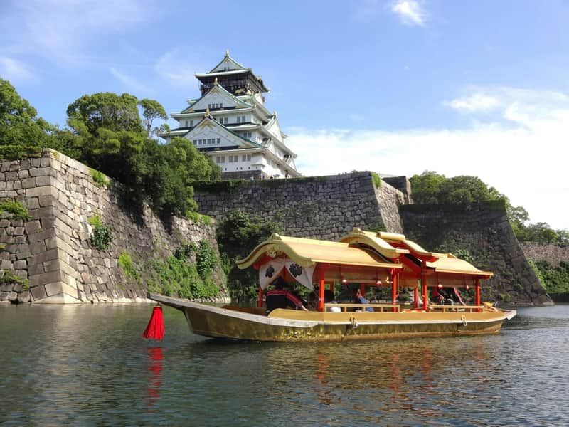 A traditional boat with a golden roof on a moat near a historic Japanese castle surrounded by stone walls and greenery.