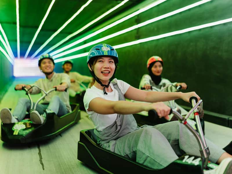 A group of people riding go-karts in a green-lit tunnel, smiling and enjoying the activity.