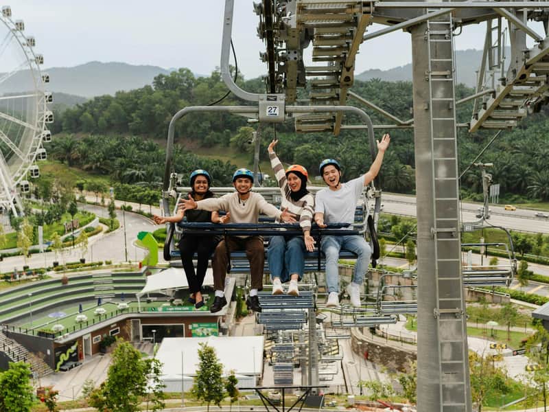 Four people riding a chairlift, smiling and waving, with a scenic background of trees and infrastructure.