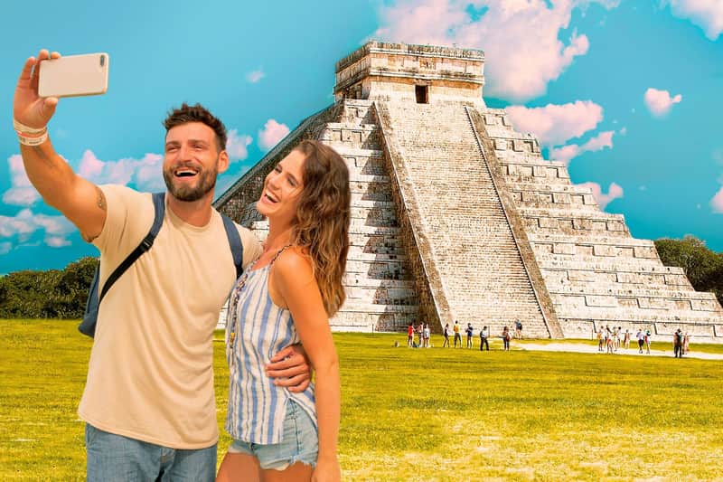 A smiling couple takes a selfie in front of an ancient pyramid on a sunny day, with a blue sky and a grassy area around.