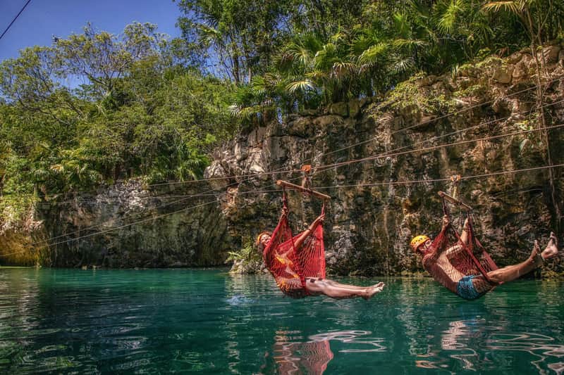 Two people ziplining over a turquoise water body in a lush, tropical setting with rocky cliffs and greenery.