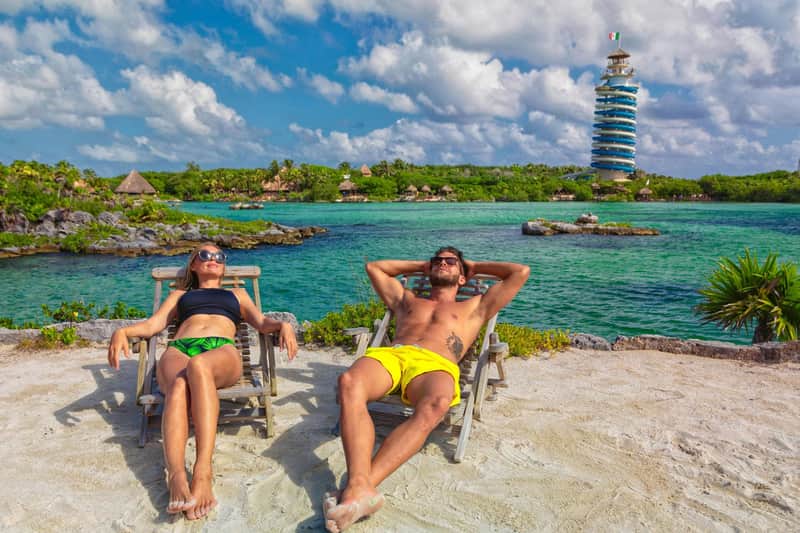 Two people relaxing on lounge chairs by a tropical waterfront with a tower and lush greenery in the background.