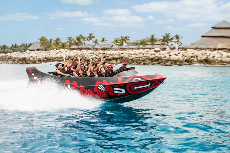 A group of people on a speeding boat decorated with red patterns, in a tropical setting with palm trees and rocks.