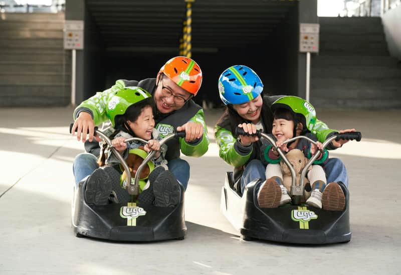 Two adults and two children wearing helmets riding luge carts indoors, smiling and enjoying the activity.