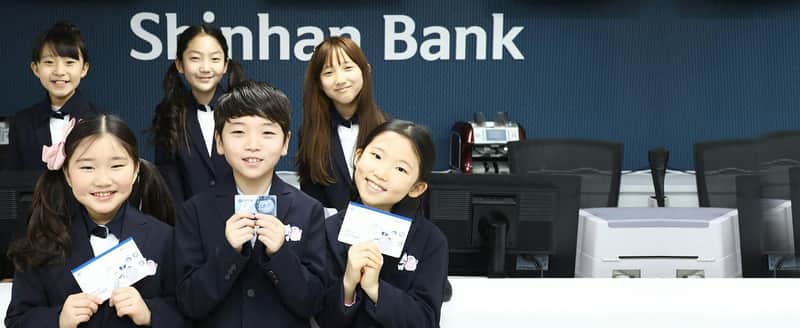 A group of children in formal attire smiling and holding cards, standing in front of a backdrop with text "Shinhan B."
