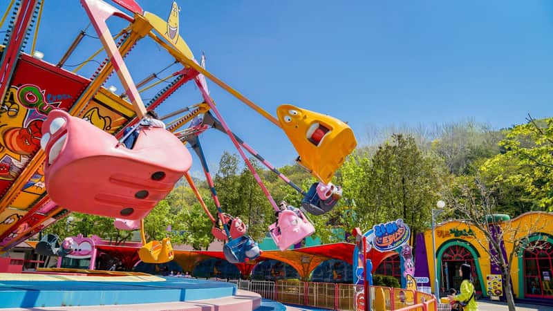 A colorful amusement park ride shaped like cartoon animals is spinning with a blue sky and trees in the background.