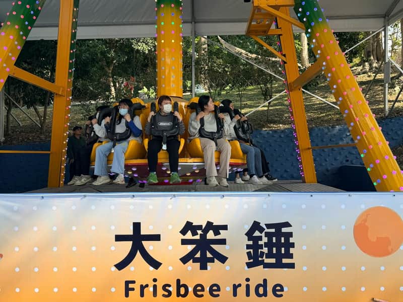 People seated on a frisbee ride at an amusement park, with a sign displaying its name in Chinese and English.