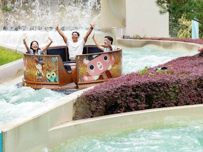 Three people on a water ride, raising their arms and smiling, with colorful cartoon graphics on the ride's side.
