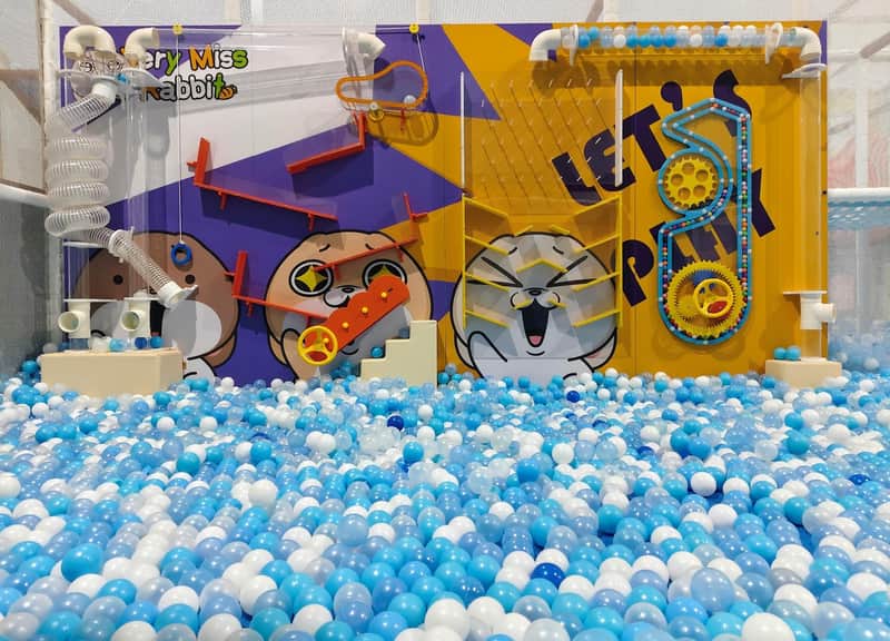 A colorful indoor play area with a ball pit, cartoon characters on the wall, and interactive elements like gears and tracks.