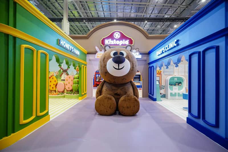 A large teddy bear statue sits in the center of a colorful indoor play area with labeled sections like "Honey's Farm" and "Baby's Clinic."