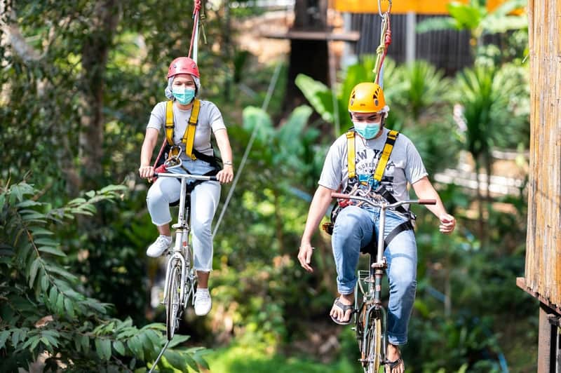 Two people wearing helmets and safety gear ride bicycles suspended on a zipline in a lush, forested setting.