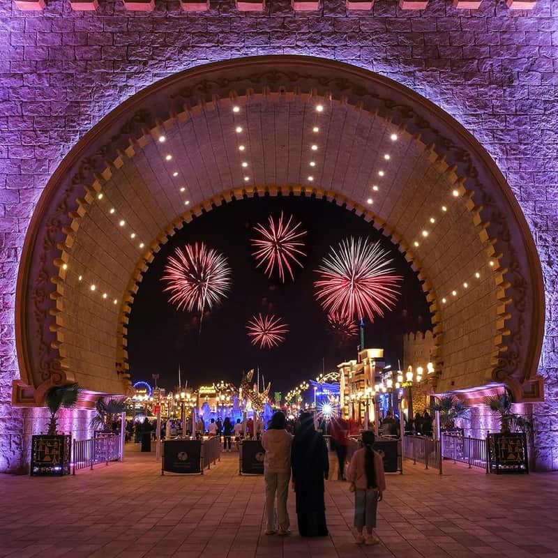 People standing under an illuminated archway, watching vibrant fireworks over a lively, well-lit amusement park at night.