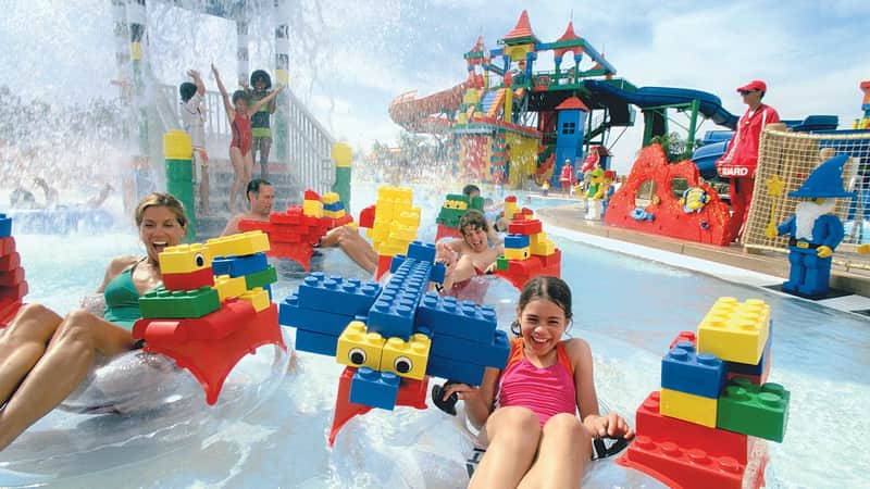 Children and adults enjoy a water park filled with large, colorful LEGO structures and slides. A girl in a pink swimsuit smiles in the foreground.