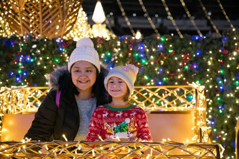 Two people wearing winter attire smiling in a festive, illuminated setting with decorative lights and greenery.