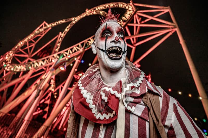A person in clown makeup and costume standing in front of an illuminated roller coaster at night.