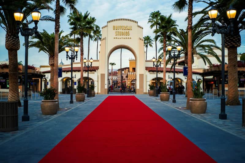 A red carpet leads to the Universal Studios Hollywood entrance arch, surrounded by palm trees and decorative lighting.