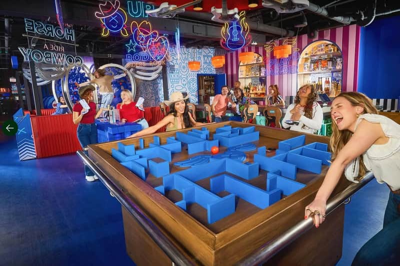 People enjoying a game with a large blue maze-like structure on a table in a vibrant indoor setting with neon signs.