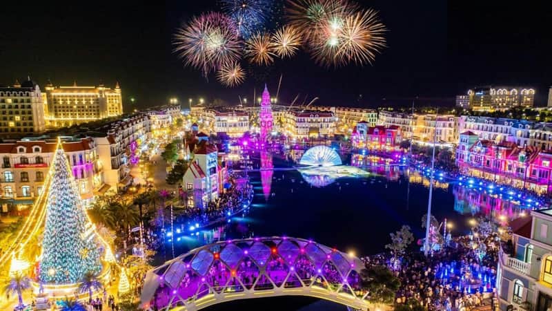 A vibrant nighttime scene with colorful fireworks, illuminated buildings, and a lit bridge over a water feature.
