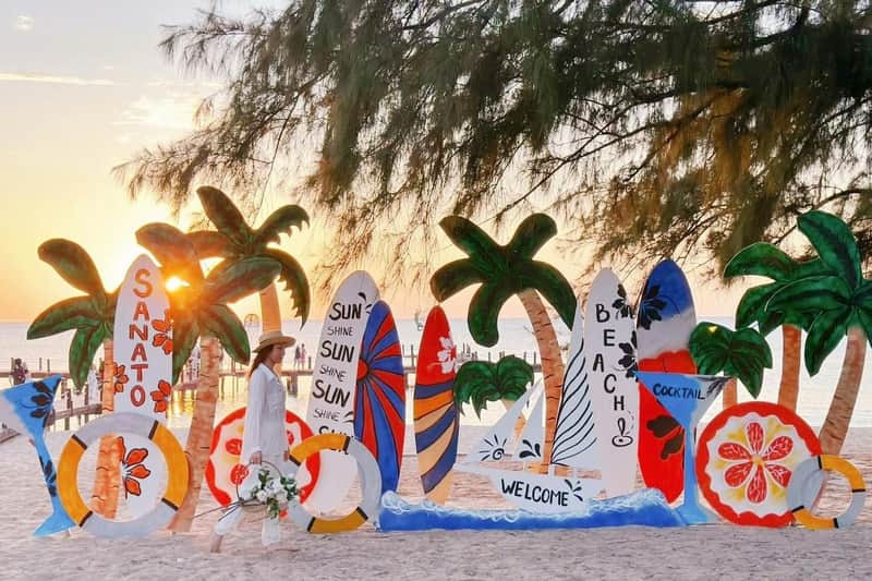 A person in white walks by colorful surfboards decorated with tropical designs on a beach, with a sunset in the background.