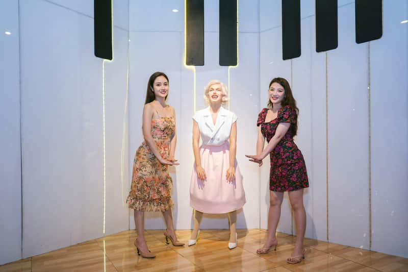 Two women pose next to a statue of a woman in a bright room with reflective flooring and wall decorations.