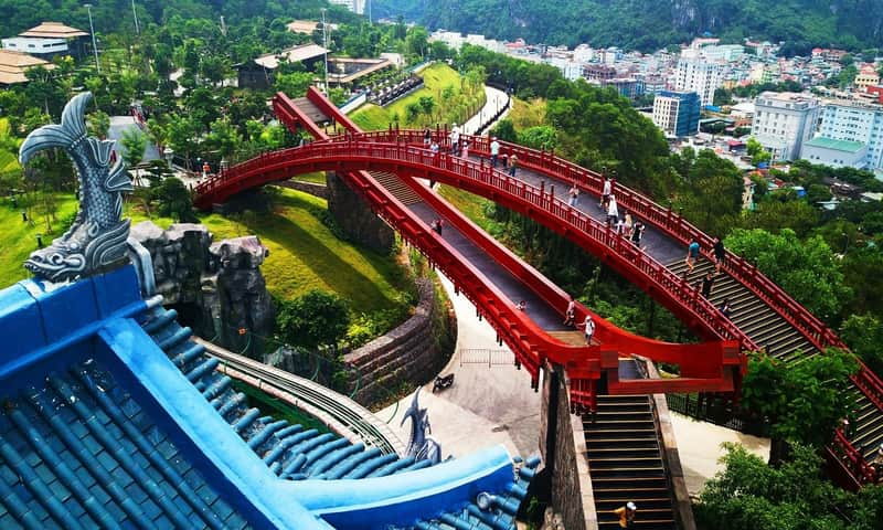 A red pedestrian bridge with decorative railings spans a lush green park. People are walking on the bridge under a bright sky.