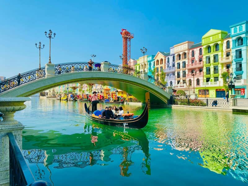 A gondola with passengers gliding through a canal under a bridge, surrounded by colorful buildings and a clear blue sky.