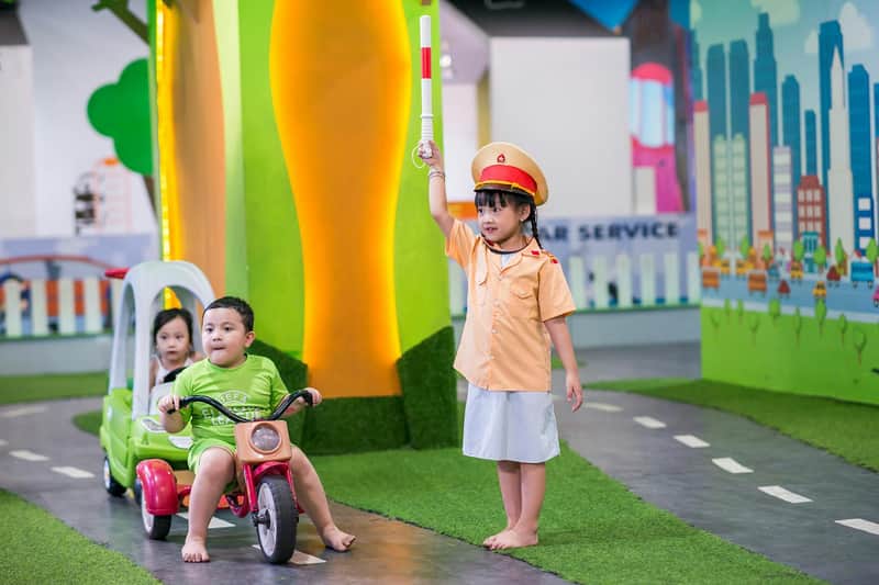 Children playing in a traffic-themed indoor area; one rides a tricycle, another sits in a toy car, and one acts as a traffic officer.