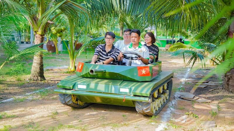 Four people riding a small green tank vehicle on a path surrounded by palm trees in a tropical setting.